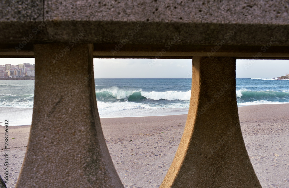 Waves breaking against a beach with sunset light. View through a handrail in a promenade. La Coruna, Spain, autumn storm.