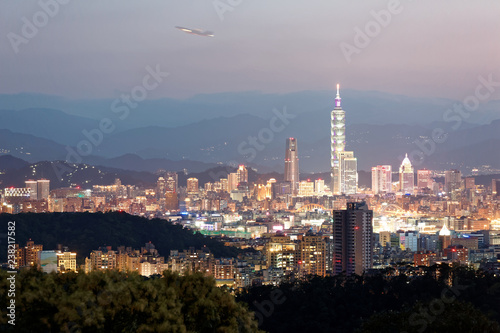 Night scenery of Taipei, the vibrant capital city of Taiwan, with landmark Tower standing amid skyscrapers, an airplane taking off from Songshan Airport & mountain silhouettes in evening twilight