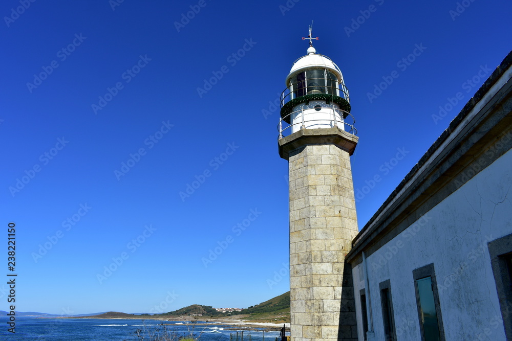 Old abandoned lighthouse with wind vane. Blue sea with waves and foam, clear sky. Sunny day, Galicia, Spain.