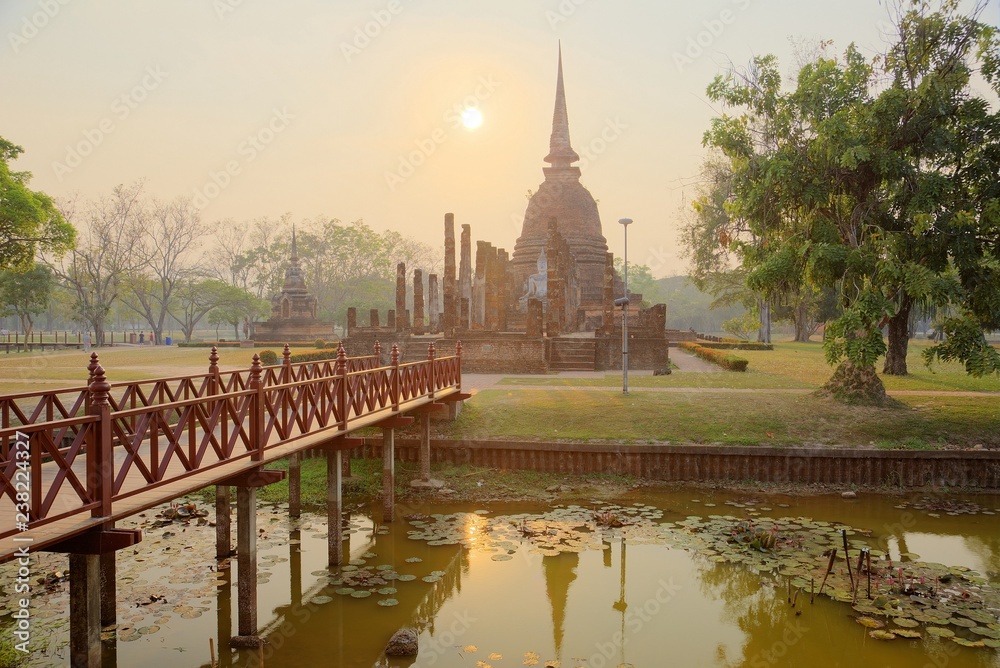 Obraz premium Sunset scenery of Wat Sa Si in Sukhothai Historical Park with the setting sun in background, a wooden bridge in foreground and the majestic temple in the middle ~ A beautiful heritage site in Thailand