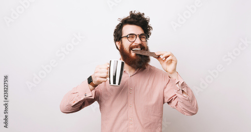 Cheerful happy bearded man in causal eating bar of chocolate and holding cup of coffee