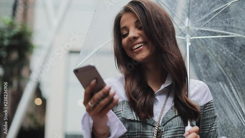 Beautiful young business woman using smartphone on the street in rainy weather, smiling, holding umbrella, communication concept photo