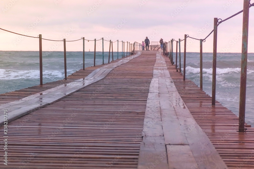 Family are walking at the sea pier in storm. Waves splashes through the wooden pier in the sea. Beautiful seascape in sunset.
