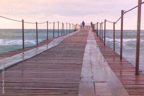 Family are walking at the sea pier in storm. Waves splashes through the wooden pier in the sea. Beautiful seascape in sunset.