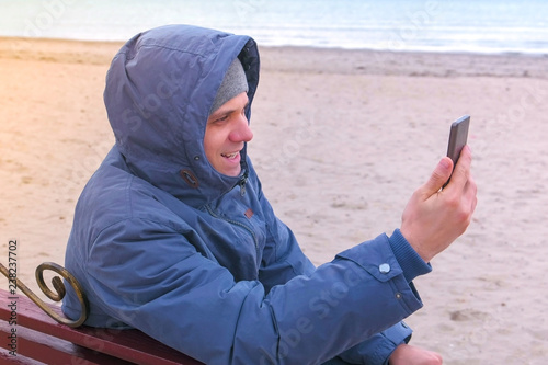 Man blogger in a blue down jacket sitting on a bench on the sand beach and talking a video chat on mobile phone.