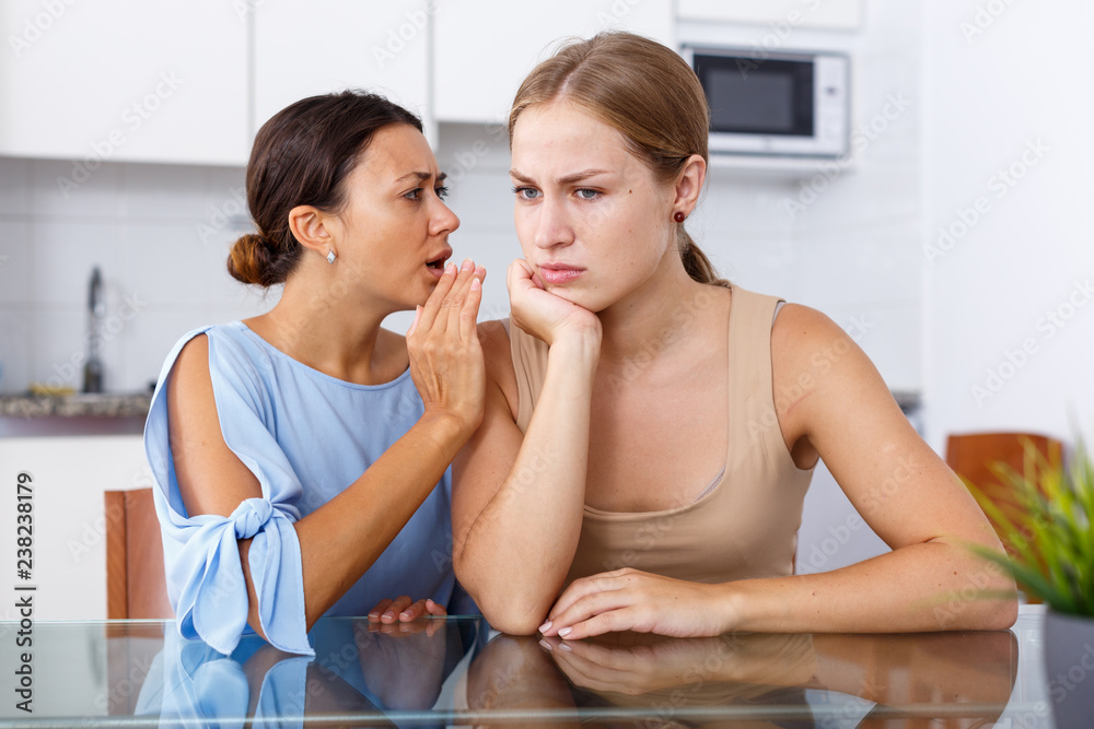 Upset young women friends whispers in the ear  and talking at table in home