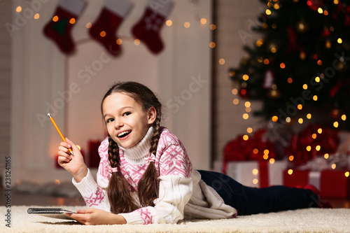 Young girl with notepad and pencil lying near christmas tree at home