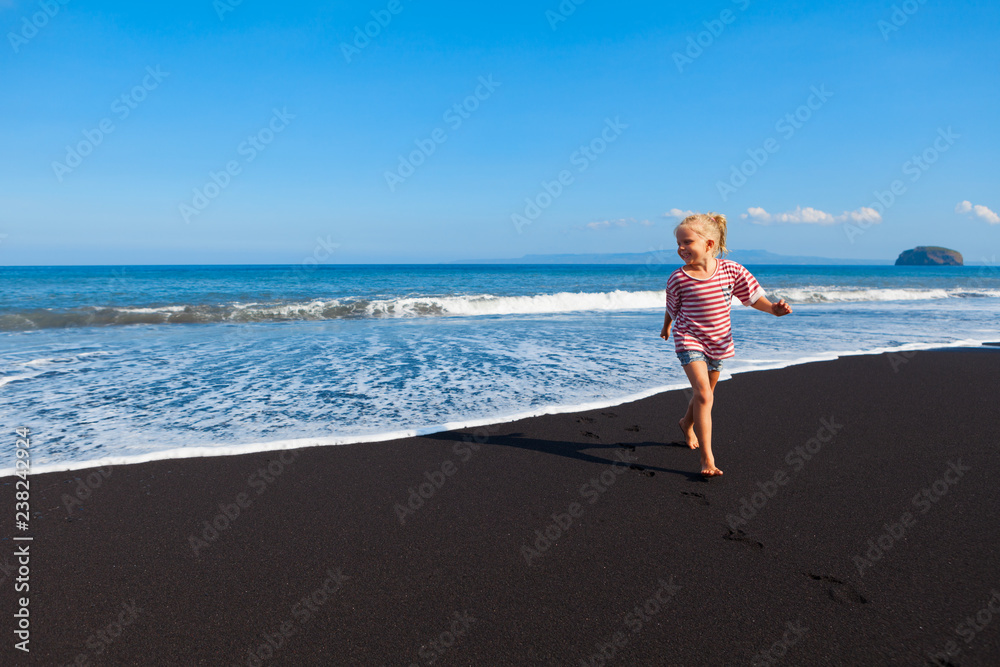 Happy barefoot kid have fun on beach walk. Run and jump by black sand along sea surf. Family travel lifestyle, outdoor sports activities, games. Summer vacation with children on tropical island Bali