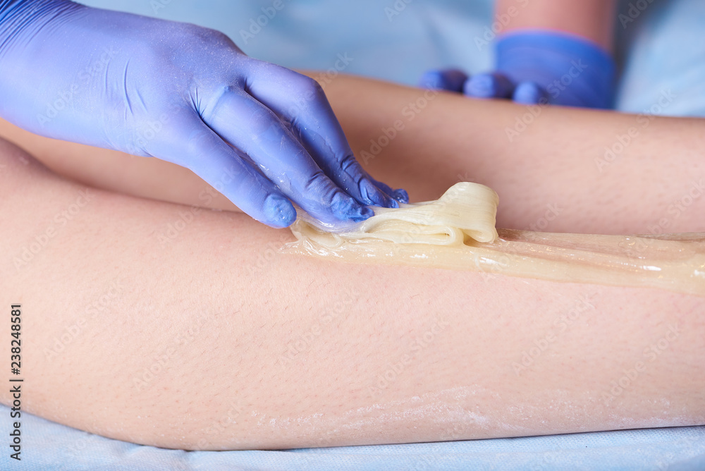 Naklejka premium Cosmetologist's hand with sugar wax doing depilation on legs