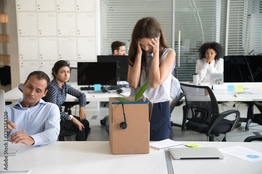 Fototapeta premium Desperate dismissed woman holding head in hands at workplace, upset female employee pack belongings in cardboard box, feeling stressed about job loss, unexpected undeserved dismissal, discrimination