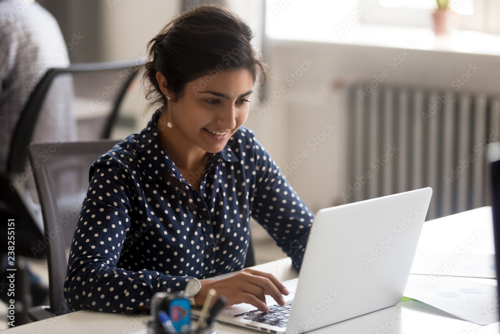 Smiling Indian female employee using laptop at workplace, looking at screen, focused businesswoman preparing economic report, working online project, cheerful intern doing computer work, typing - obrazy, fototapety, plakaty 