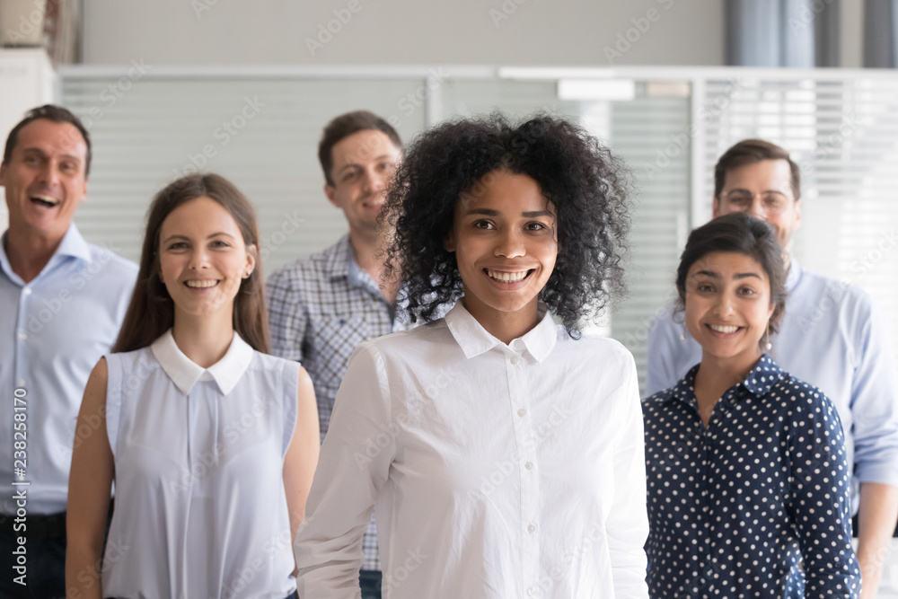African American leader, coach posing, standing with multiracial group of employees in office, diverse happy group team staff members at background, successful business owner, look at camera
