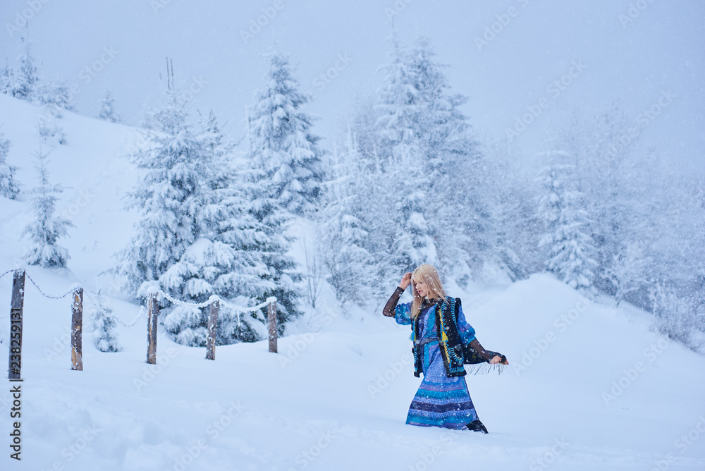 Attractive fashionable slim blond girl in long blue dress, embroidered warm sleeveless fur coat outdoor running in deep clean snow on bright sunny winter day on blurred fir-trees background.