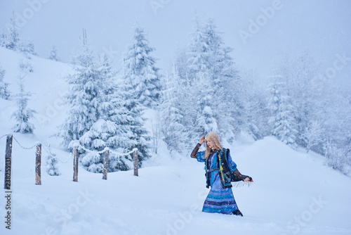 Attractive fashionable slim blond girl in long blue dress, embroidered warm sleeveless fur coat outdoor running in deep clean snow on bright sunny winter day on blurred fir-trees background. © anatoliy_gleb