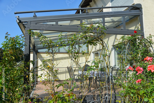 Verglaster Wintergarten mit beschichtetem Metallrahmen, Windmesser und Glasdach, umgeben von Rosenbögen an einem Wohnhaus