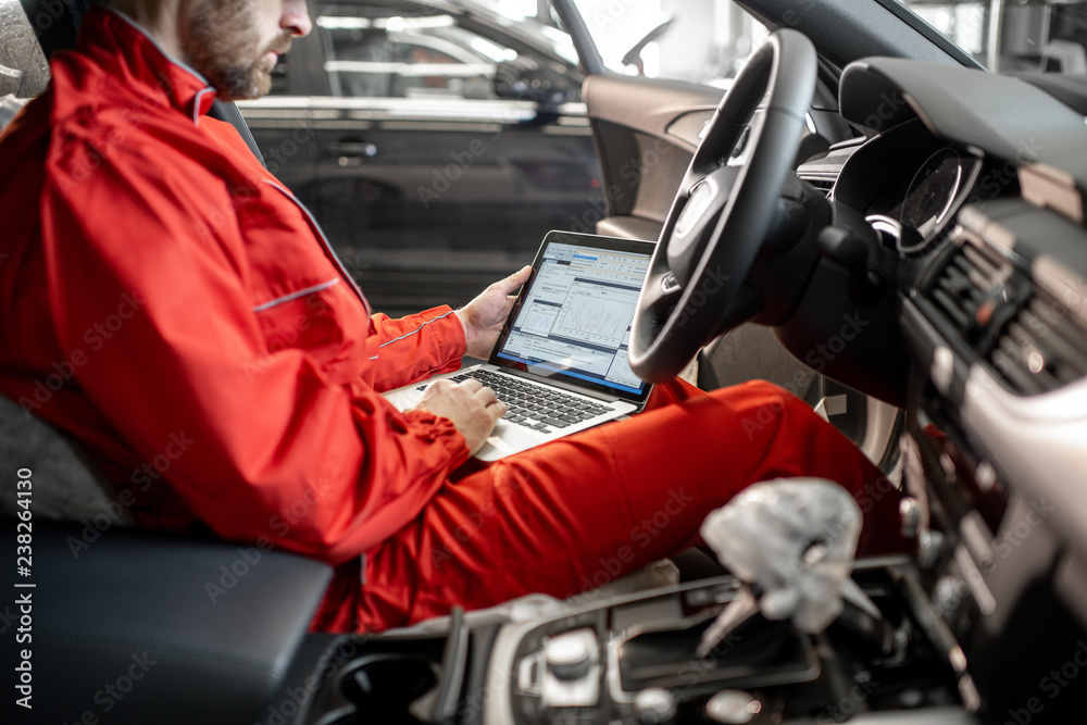 Auto mechanic in red uniform diagnosing car with computer sitting on the driver seat at the car service