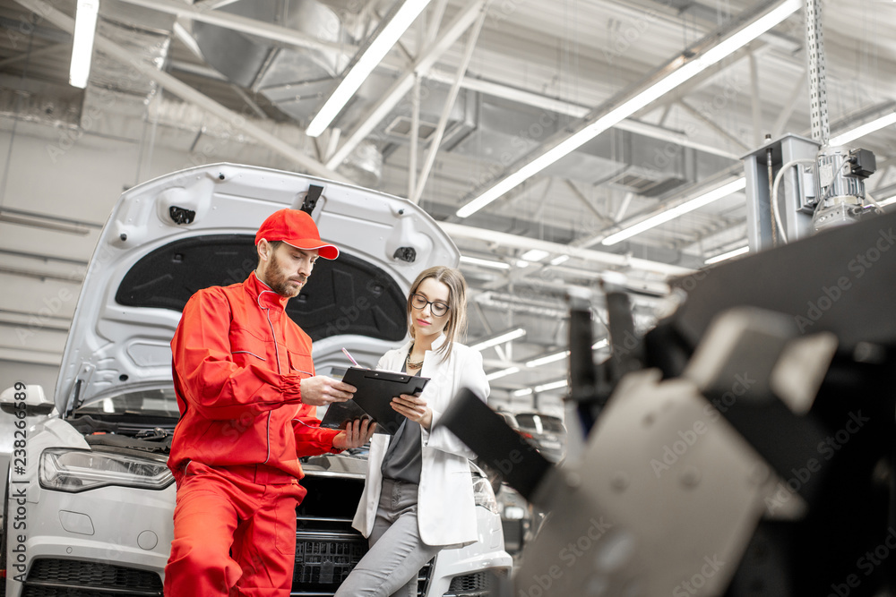 Young woman client with auto mechanic in red uniform standing with some documents at the car service. Wide view with copy space