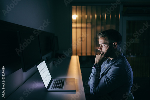 Young concerned bearded Caucasian businessman looking at laptop and finding a solution for the problem while sitting late at night in the office.