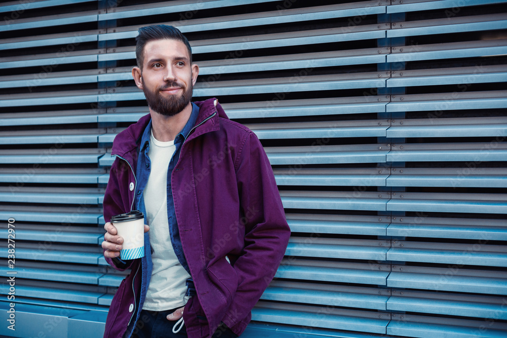 Handsome young man in purple winter jacket drinking coffee. The guy drinking coffee on the street. Handsome young man in purple winter jacket drinking coffee. The guy drinking coffee on the street.