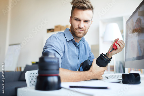 Portrait of handsome young man repairing photo camera lens sitting at desk , copy space