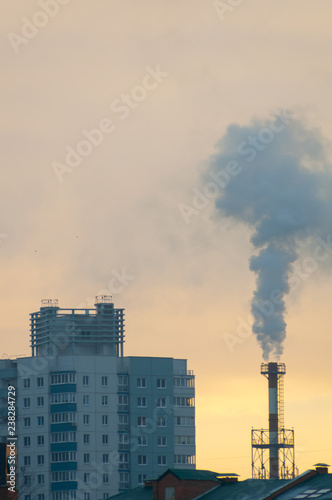 Thick smoke rises from the chimney high above the city against orange sky. City, air and environmental pollution. Climate Change Theme. View of pipe with smoke and residential apartment buildings.