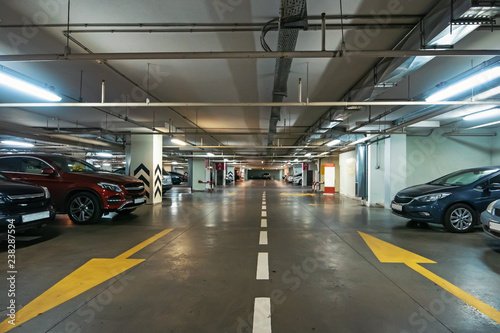 Illuminated underground car parking interior under modern mall with lots of vehicles and arrows on floor © DedMityay