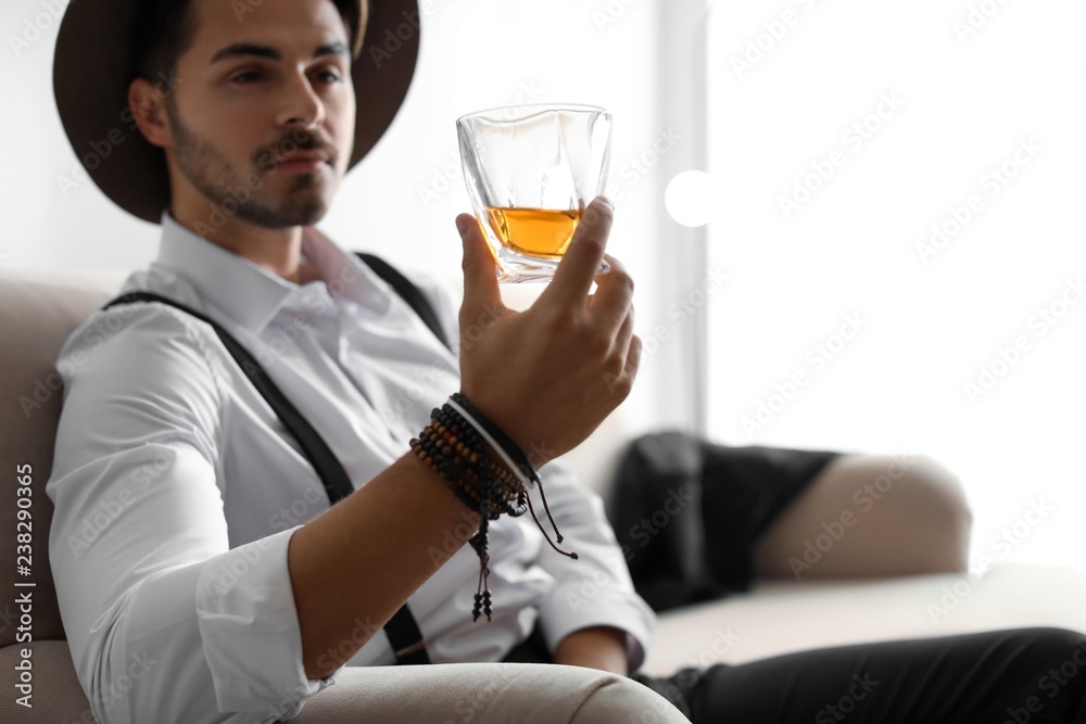 Young man with glass of whiskey sitting on sofa at home. Space for text