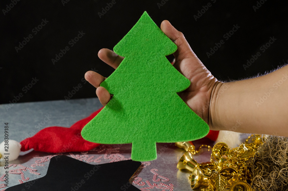 Man hand holding a christmas tree against christmas decoration. Christmas decoration with a christmas tree on the center, Santa Claus hat, star shape chalkboard and golden bells