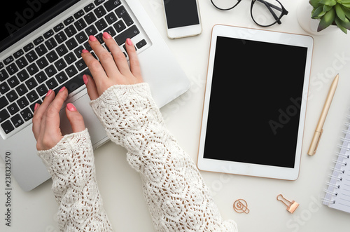 Top view of woman's hands typing on laptop keypad placed on white office desktop with blank smartphone, decorative plant and supplies. Mock up. Technology devices view from above. Office equipment.