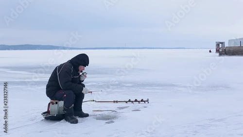 Elderly fisherman in dark clothes fishing on winter fishing rod on frozen river and drink hot tea or coffee. Winter ice fishing.