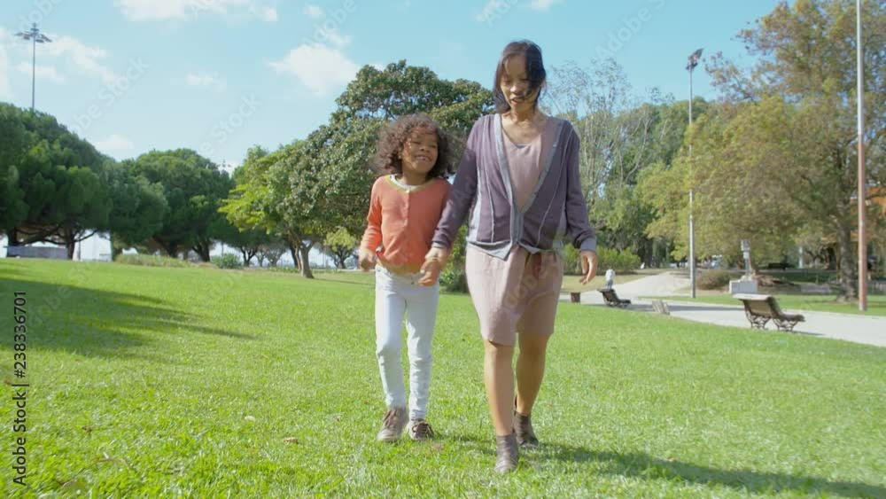 African American daughter holding hands with Asian mother and walking in park. Front view of mom and girl walking on green lawn in slow motion. Happy family concept African American daughter holding hands with Asian mother and walking in park. Front view of mom and girl walking on green lawn in slow motion. Happy family concept