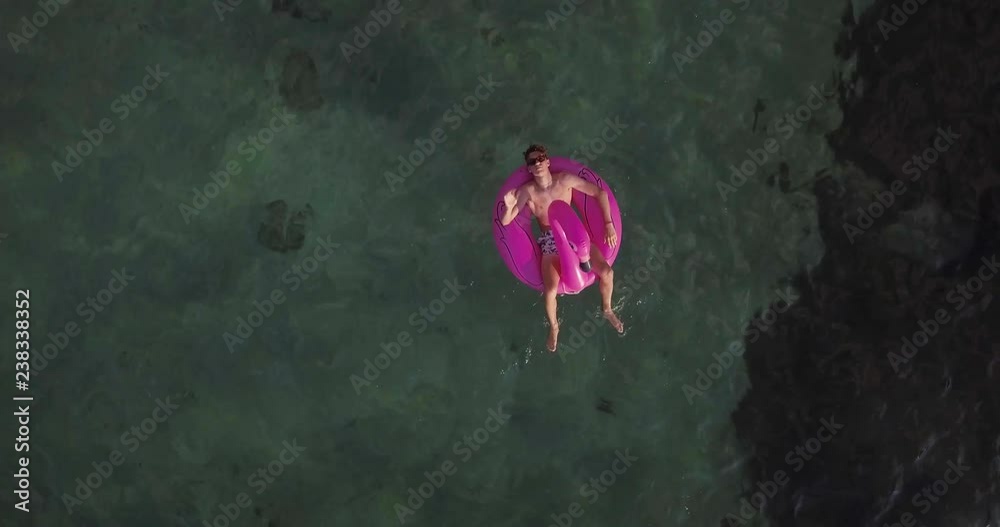 Aerial, a young man on a flamingo shaped inflatable ride on pool toy floating in the middle of the emerald sea