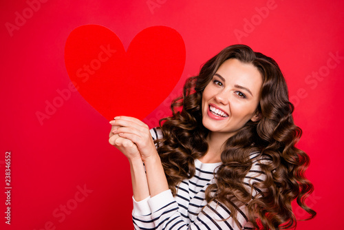 Close-up portrait of nice attractive cheerful funny wavy-haired lady wearing striped pullover holding in hands large paper heart isolated over bright vivid shine red background