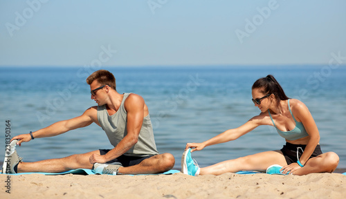 fitness, sport and lifestyle concept - happy couple stretching legs on beach before training