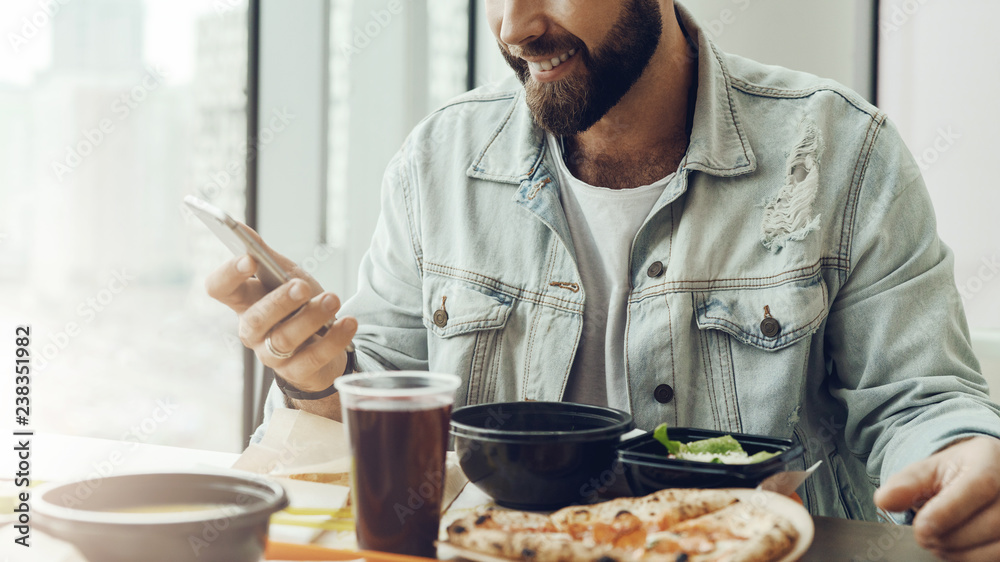 Hipster guy sits in cafe at table, has lunch, using smartphone. Man pays order in restaurant using mobile application.