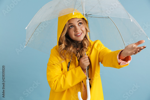 Image of pleased woman 20s wearing yellow raincoat standing under transparent umbrella, isolated over blue background