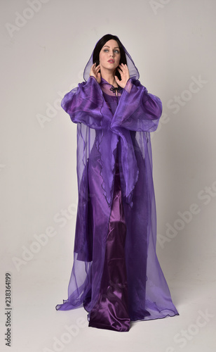 full length portrait of beautiful girl with long black hair,   wearing purple fantasy medieval gown and cloak. standing pose on grey studio background.