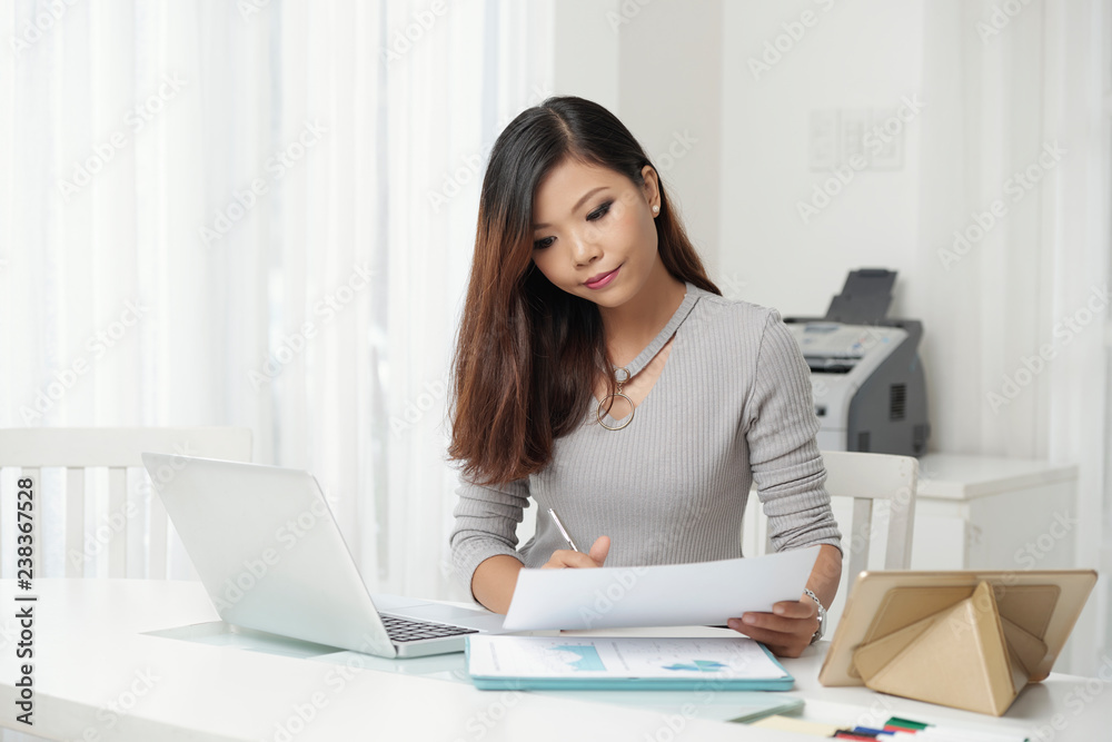 Beautiful Asian woman with papers and gadgets sitting at table in light office Beautiful Asian woman with papers and gadgets sitting at table in light office