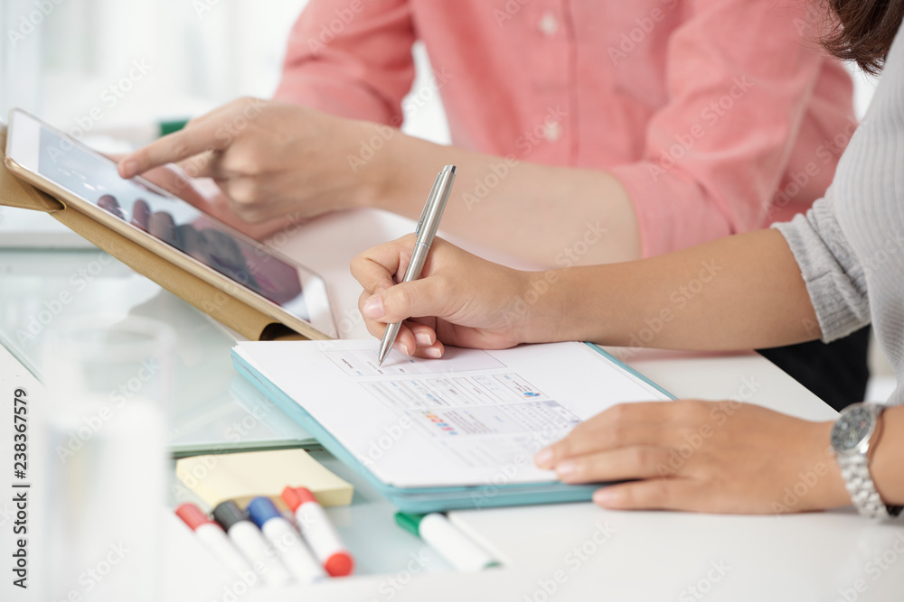 Faceless shot of contemporary colleagues using tablet while writing on clipboard sitting at table