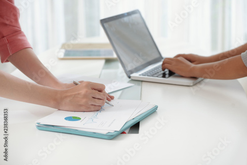 Faceless side view of man writing on paper with graph while colleague using laptop at desk