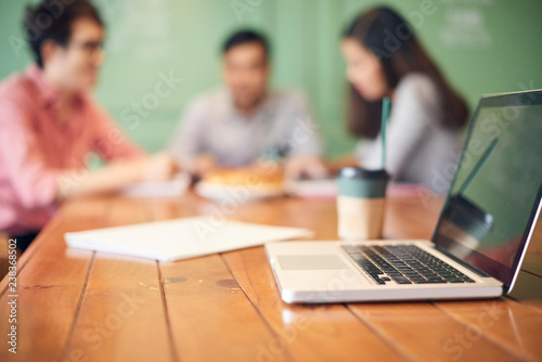 Wooden table with laptop and modern colleagues having coffee and working on background