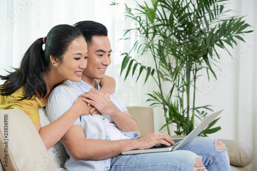 Side view of young Asian couple surfing laptop while sitting on couch at home