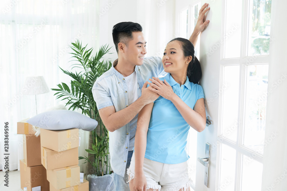 Cheerful modern Asian couple standing near window in new flat happy with moving and looking at each other Cheerful modern Asian couple standing near window in new flat happy with moving and looking at each other