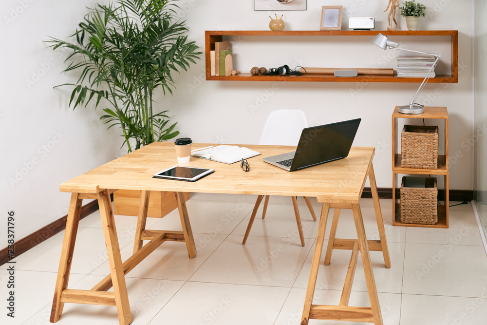 Empty wooden office table with laptop, touchpad and notepad on it with green plant and big shelf in the background Empty wooden office table with laptop, touchpad and notepad on it with green plant and big shelf in the background