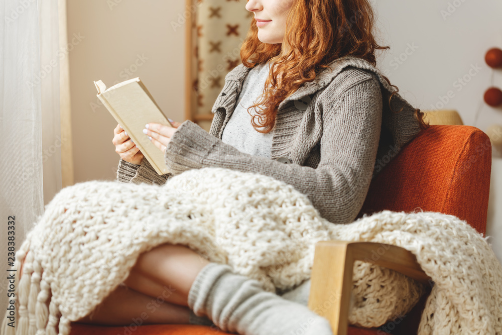 Woman with blanket on knees sitting on armchair and reading book - obrazy, fototapety, plakaty 