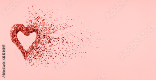 Glitter heart dissolving into pieces on pink background.  Valentines day, broken heart and love emergence concept . Living coral theme - color of the year 2019