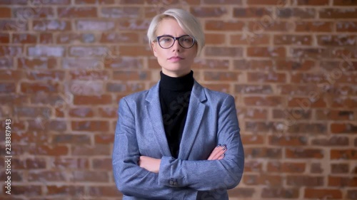 Portrait of middle-aged short-haired female in glasses watching seriously and modestly into camera on bricken wall background. photo