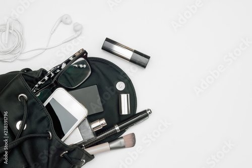 Top view of woman black bag open out with accessories smartphone, perfume, pens, cosmetics, earphone and glasses  on white background