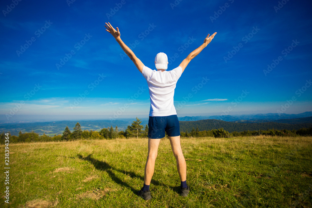 Summer hiking in mountains. Young tourist man in cap with hands up on top of mountains admires nature. Summer hiking in mountains. Young tourist man in cap with hands up on top of mountains admires nature.