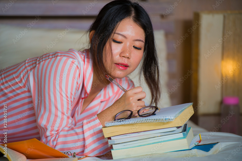 smart Asian American student girl at home bedroom sitting on bed studying for exam with laptop computer and textbooks focused and concentrated in education concept smart Asian American student girl at home bedroom sitting on bed studying for exam with laptop computer and textbooks focused and concentrated in education concept